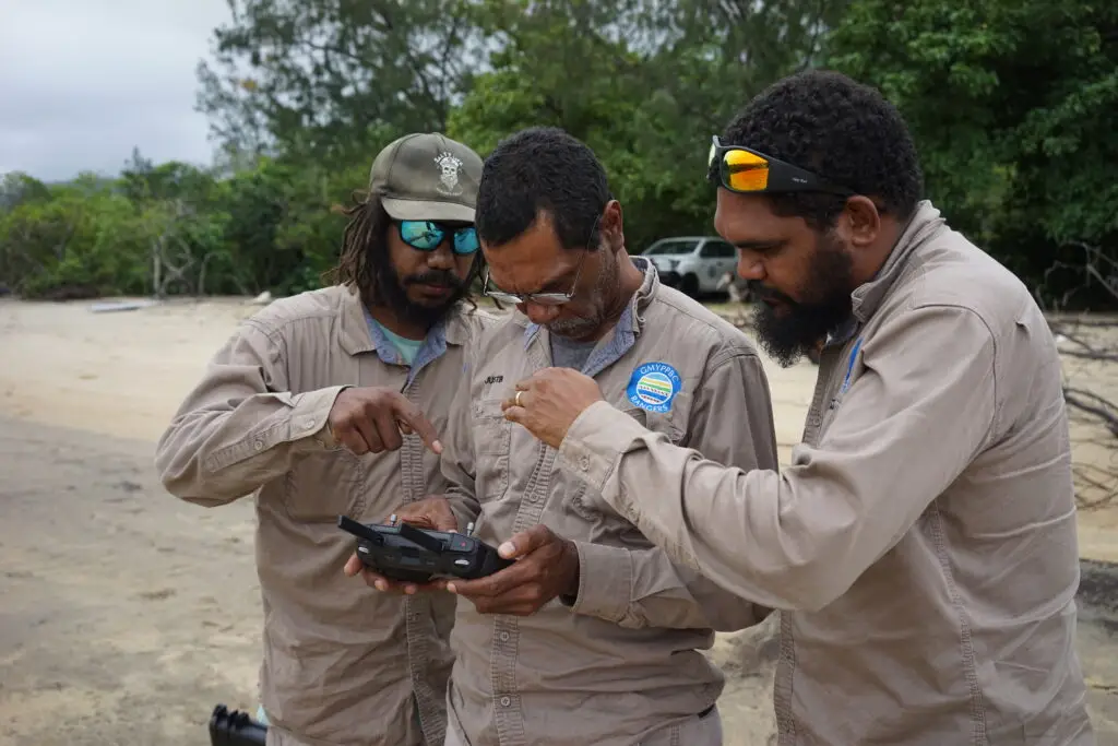 Three people discussing drone controls on beach.